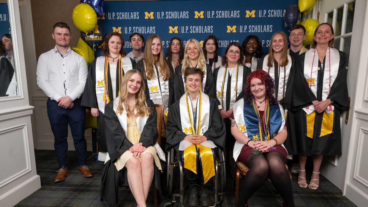 3 rows of University of Michigan students, wearing graduation stoles and cords, pose before a U.P. Scholars backdrop