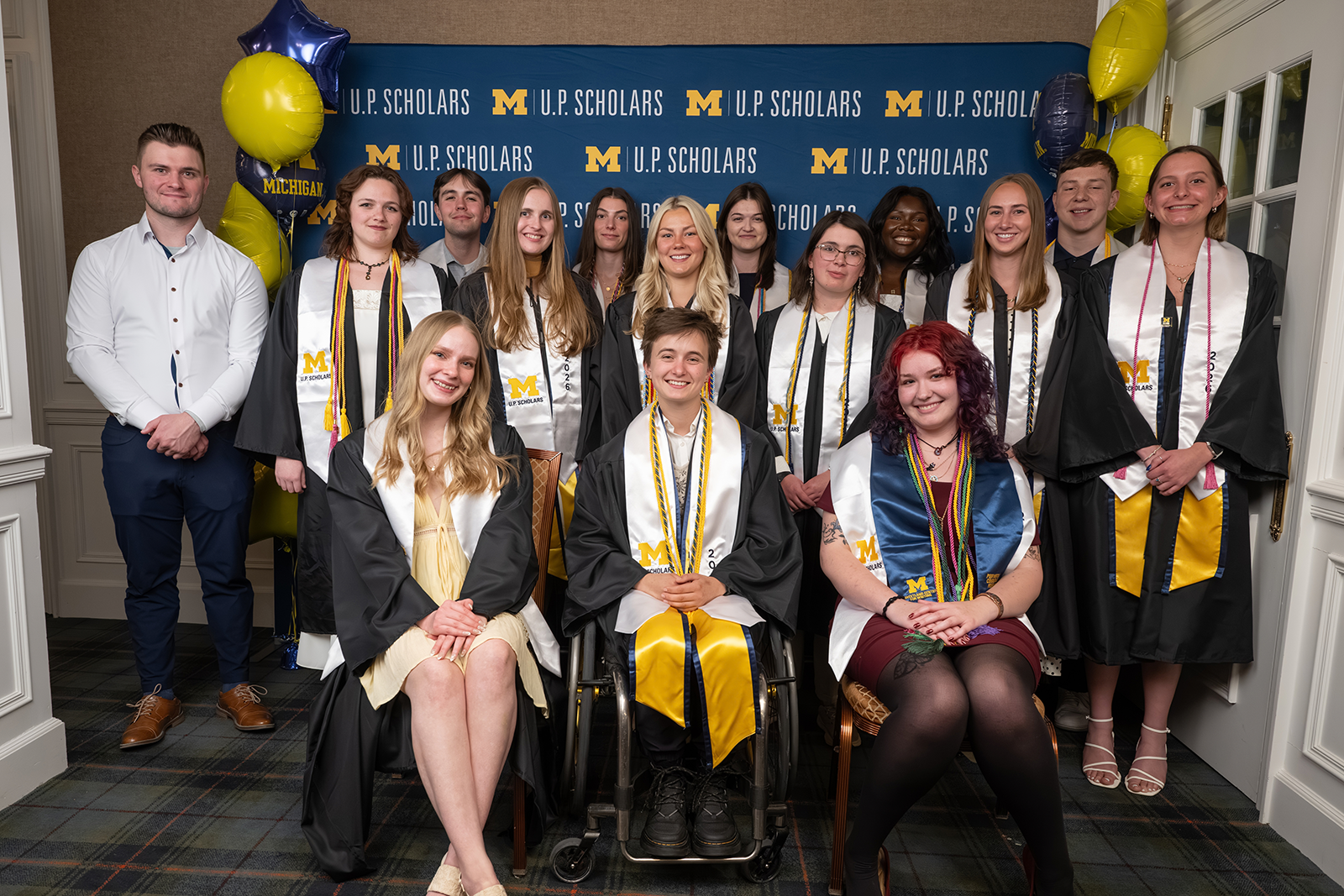 3 rows of University of Michigan students, wearing graduation stoles and cords, pose before a U.P. Scholars backdrop
