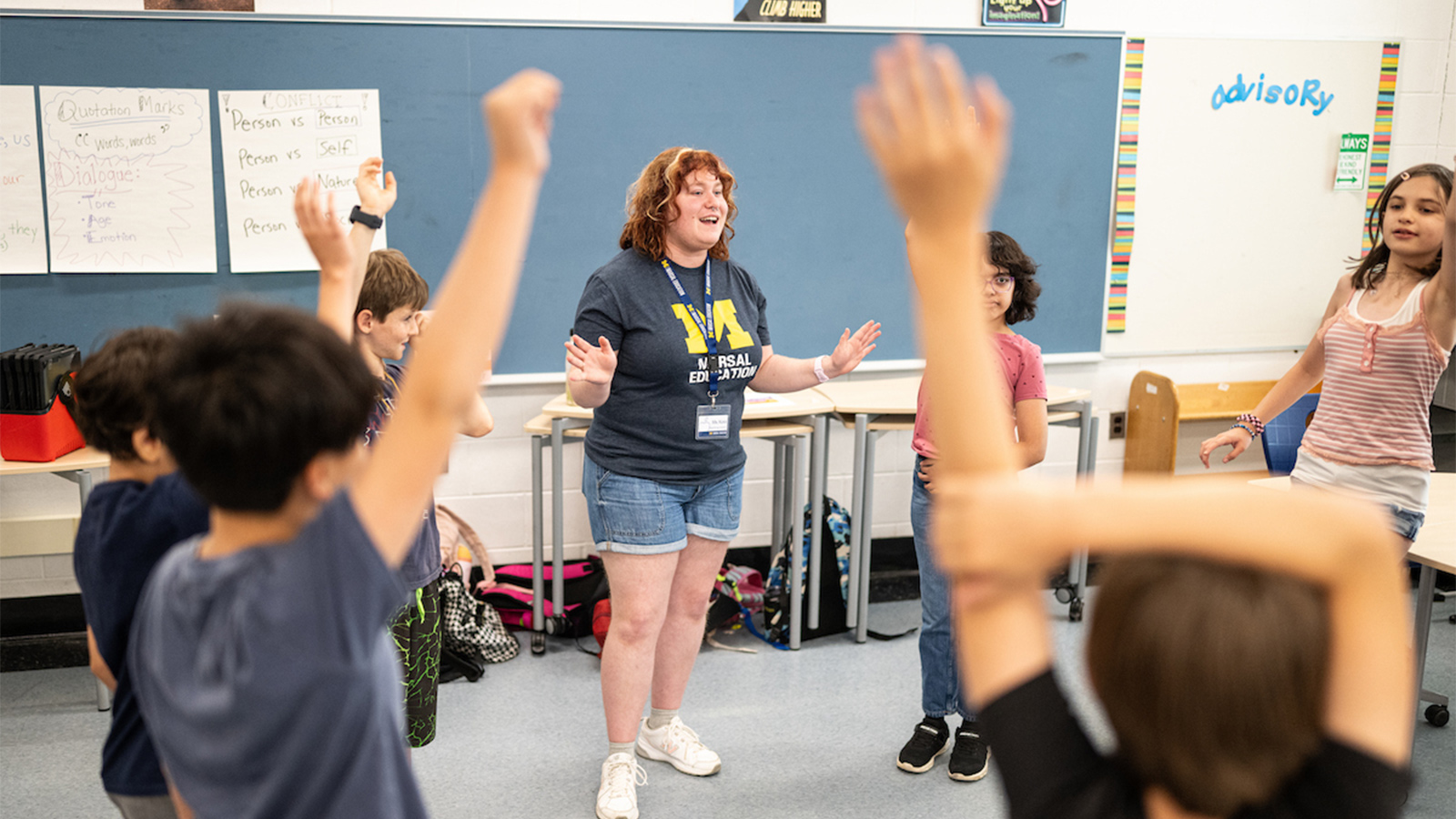 A teaching intern smiles and gestures while leading an energetic classroom activity as students gather around with raised hands.