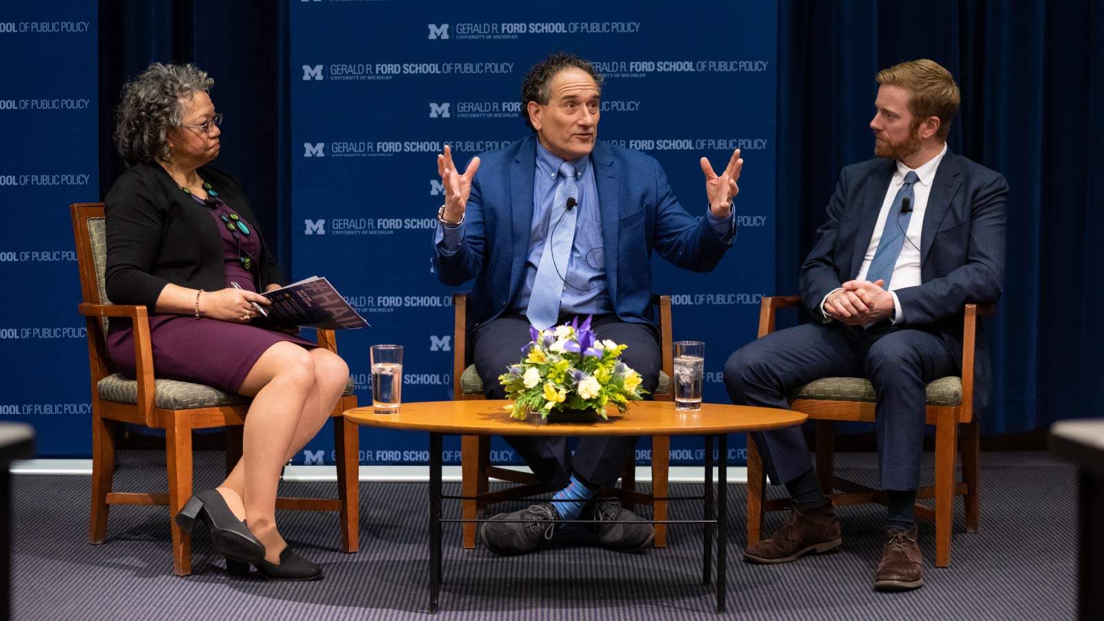 A woman and two men sitting in chairs around a coffee table having a discussion in front of a backdrop that reads “Gerald R. Ford School of Public Policy.”