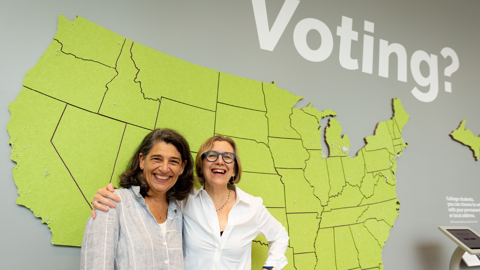  Two women standing and smiling in front of a lime green-colored map of the United States.