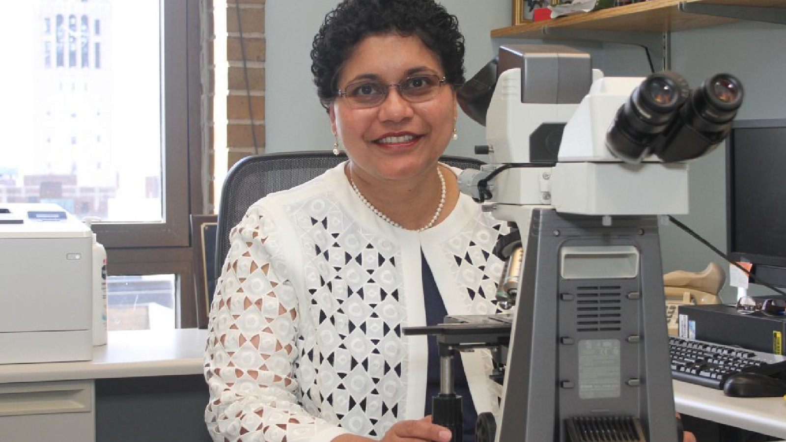 Dr. Nisha D’Silva sitting in a chair and posing by a microscope.