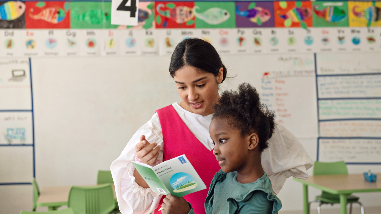 An elementary school teacher helping a young girl to read a book.