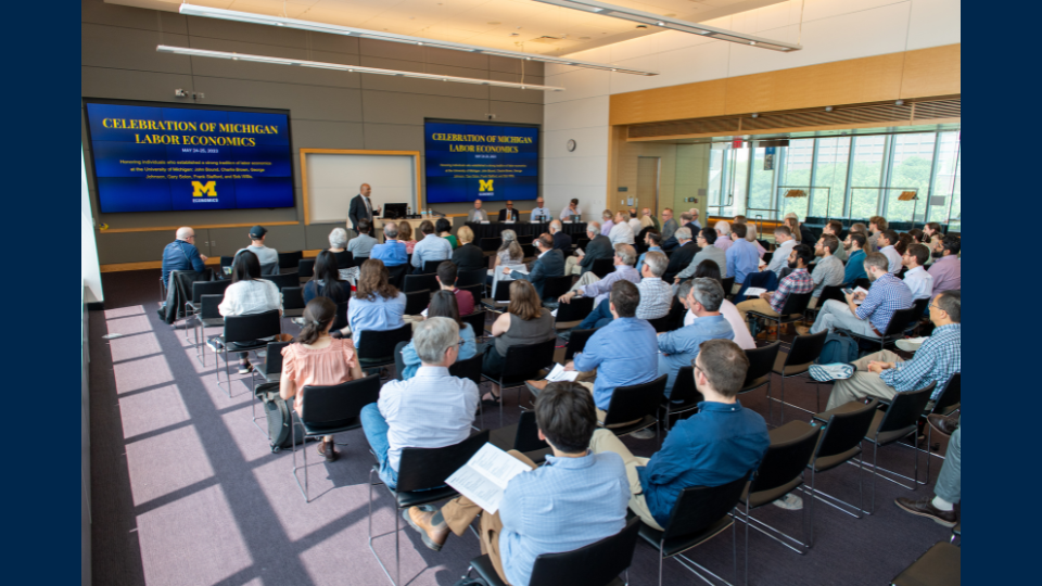 People sit in rows of chairs while watching an economics presentation