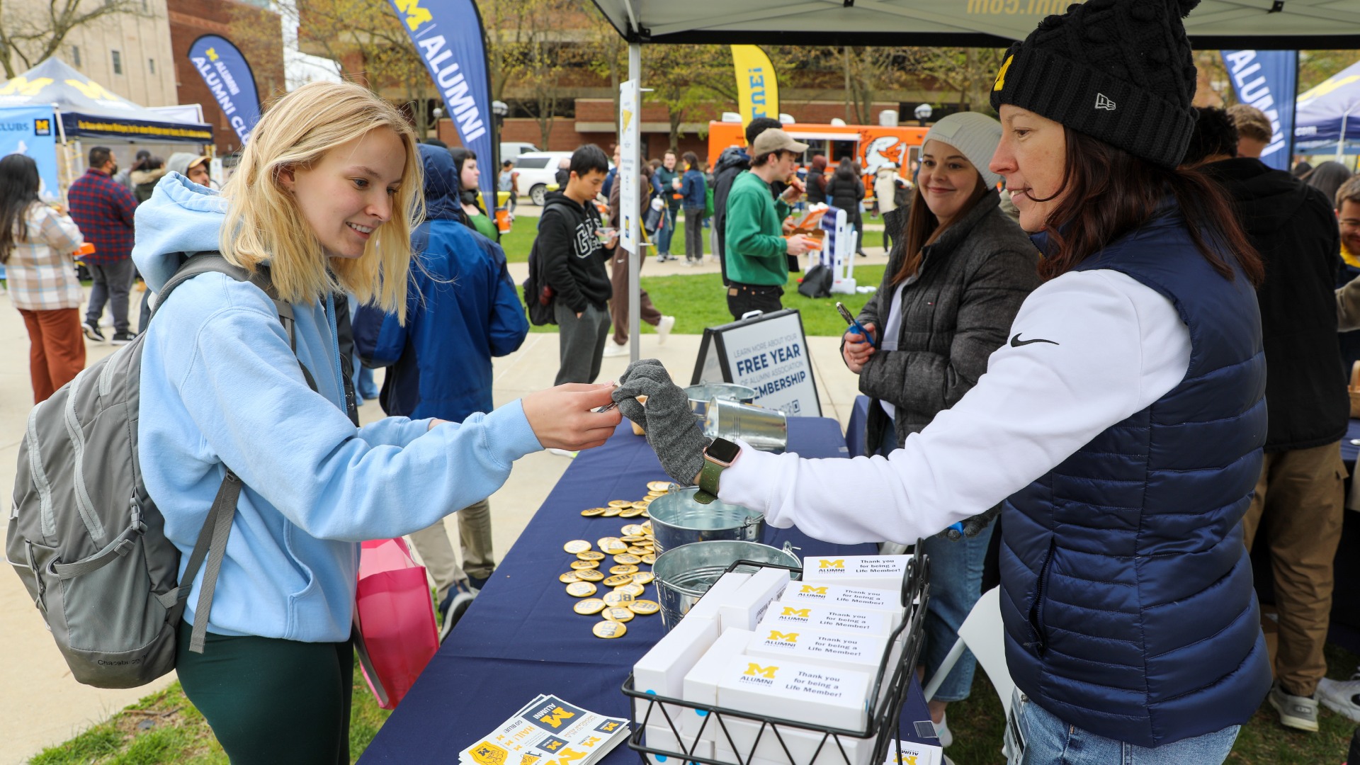 A woman approaches a booth giving away U-M swag on campus. A crowd of people is gathered in the background, where there are signs with the block M that say “Alumni” on them, and there is also a Little Ceasar’s pizza truck.
