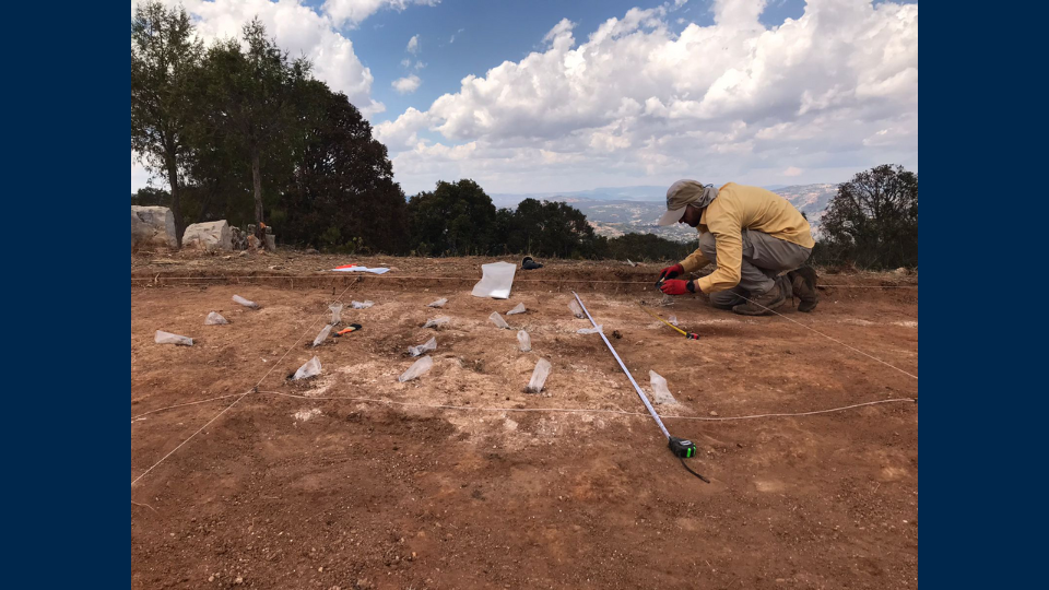 A man crouches to inspect findings uncovered in the ground at an archaeological site.