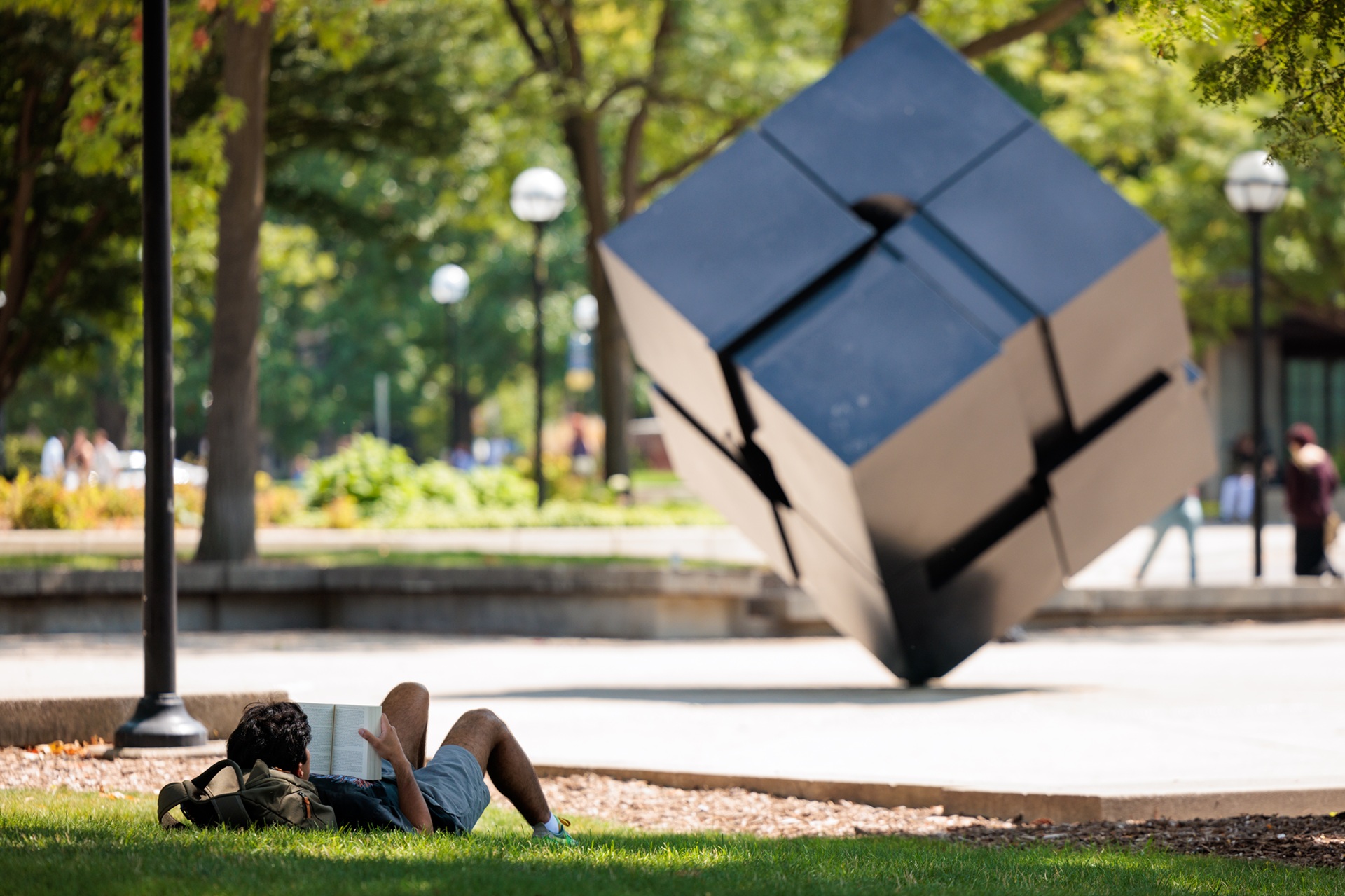 A student reclined on the grass and reading in front of the sculpture, Endover, popularly known as The Cube.