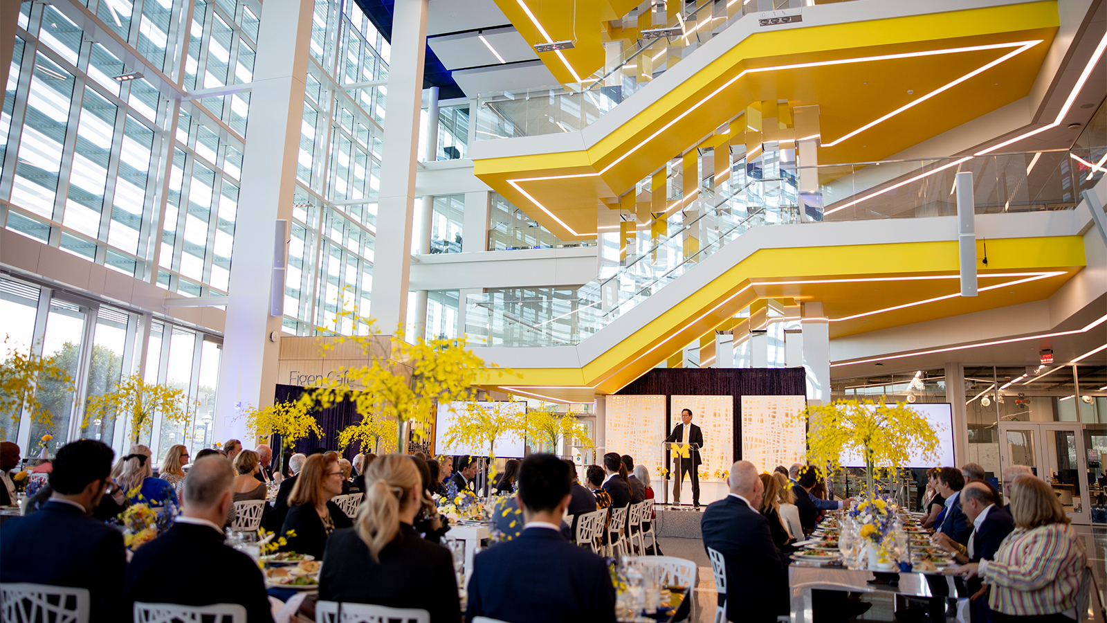 Santa Ono on stage addressing guests seated at tables in the U-M’s Ford Robotics building atrium.