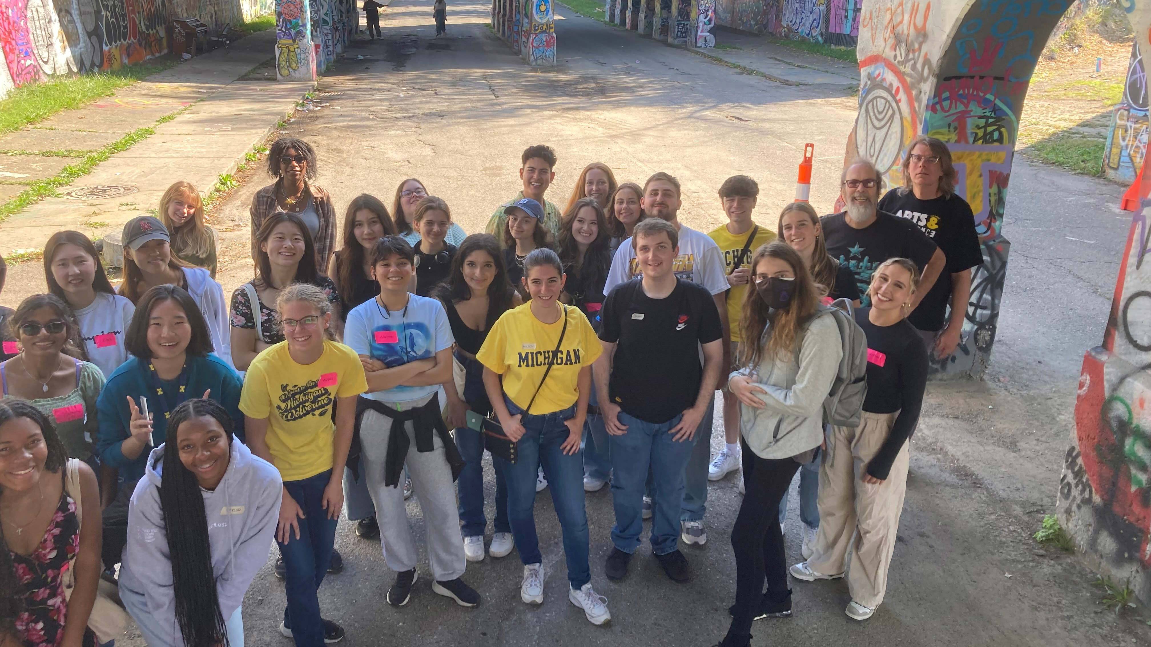 A group of about 30 casually dressed LSWA students and one instructor with gray hair and a beard, pose for a photo in front a colorful, graffiti-covered highway underpass in Detroit. They are smiling.