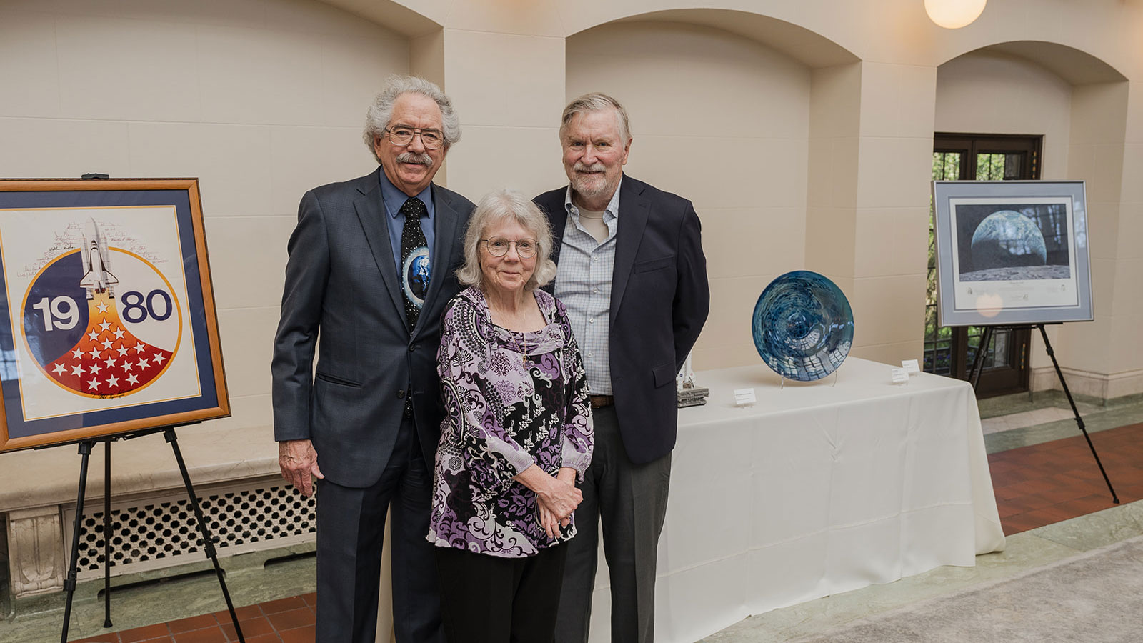 Two men and a woman stand in front of a display of artwork about space.