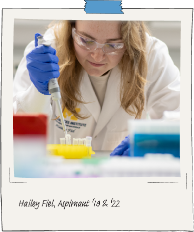 Aspirnaut student Hailey Fiel, wearing a white lab coat and blue, latex gloves leans over some test tubes in a laboratory