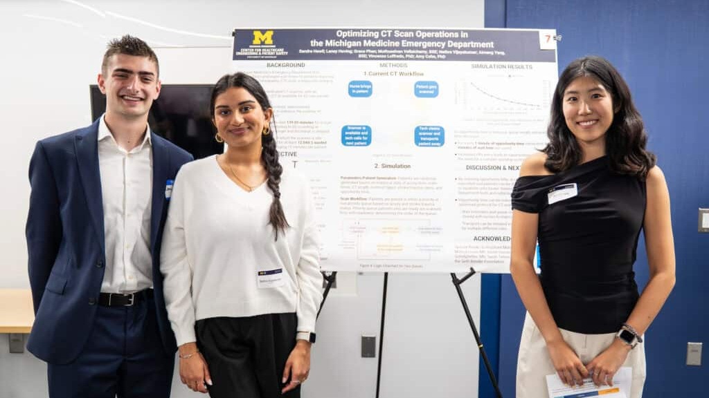Three individuals presenting a scientific poster titled "Optimizing CT Scan Operations in the Michigan Medicine Emergency Department." All are standing, one is holding a document, and are smiling and located indoors.