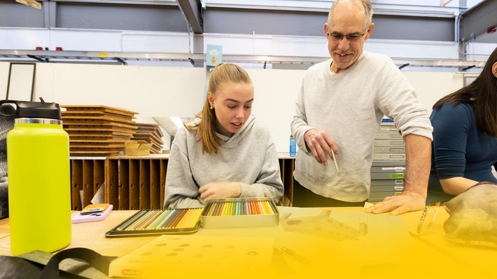 A University of Michigan student and professor are engaged in conversation at a table surrounded by various art supplies.