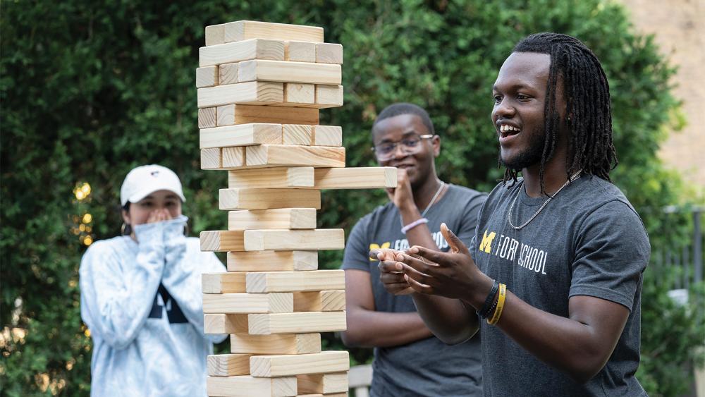 Three students outside wearing Michigan gear smiling and laughing as they play a game of giant Jenga.