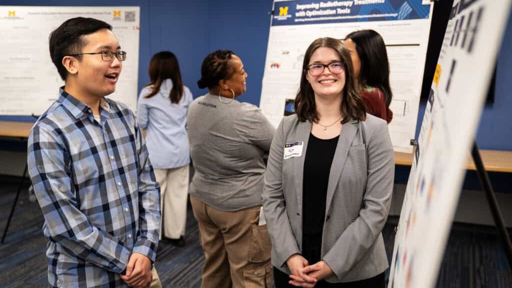 Two individuals smiling while standing in front of informational posters at a U‑M IOE event. Several other participants are engaged in discussions in the background.