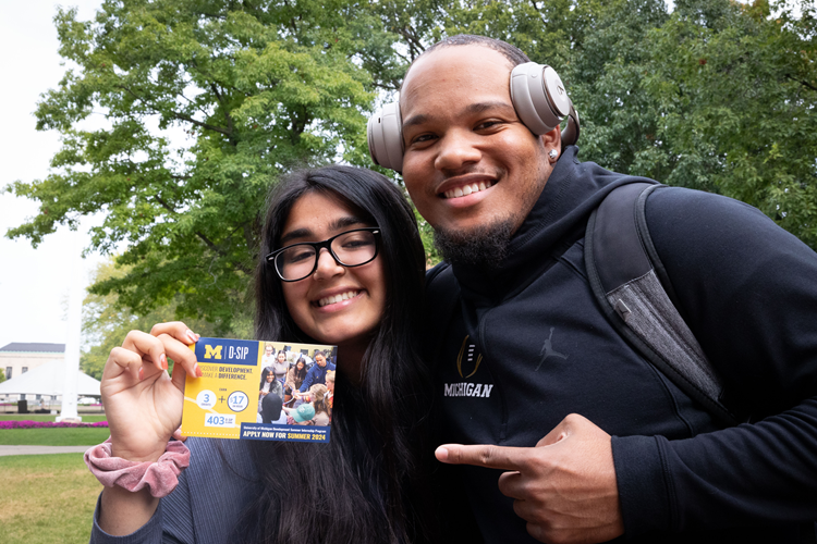 Two smiling U-M students holding up a postcard that promotes the Development Summer Internship Program.