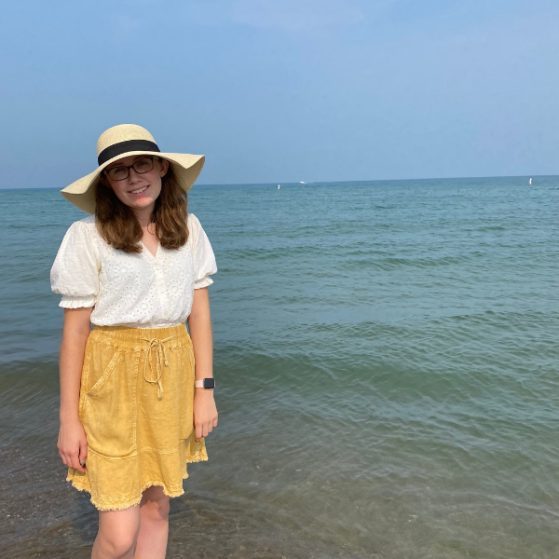 Paige Bost wearing a sun hat, white blouse, and yellow skirt stands in ankle deep water in a lake