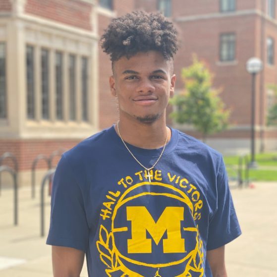 Myles Ware, wearing a U‑M t-shirt, poses in front of some brick buildings on campus