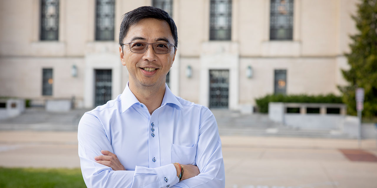 A man stands with arms crossed in front of a large stone university building, dressed in a light blue shirt.