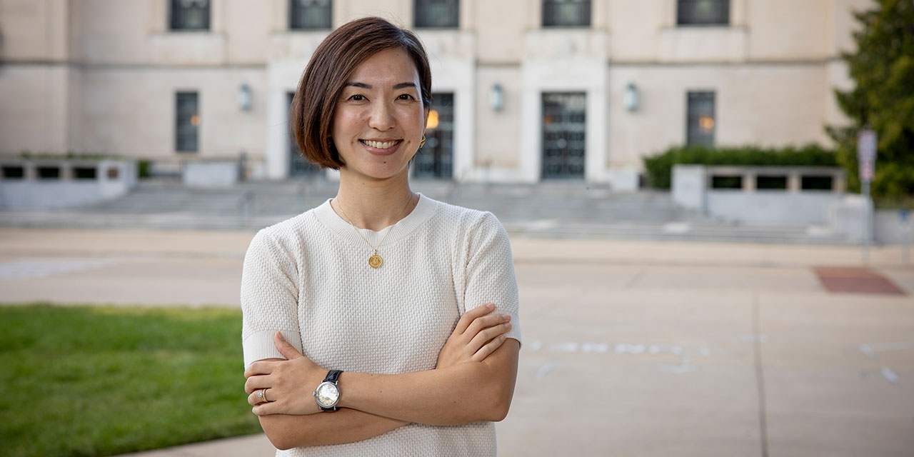 A woman stands in front of a building with crossed arms wearing a tan short sleeve sweater. 