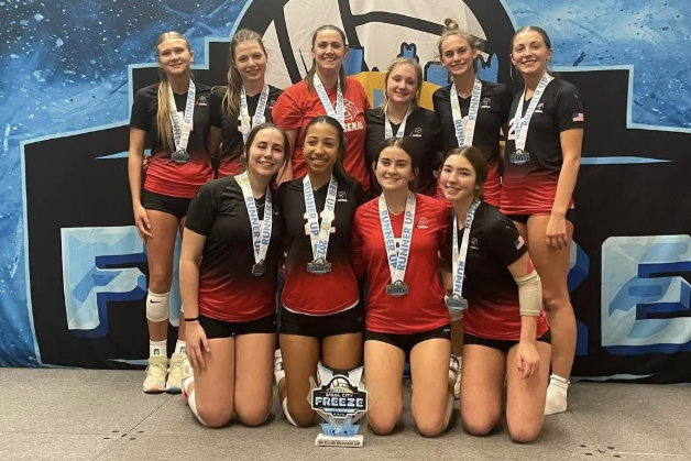 A women's volleyball team poses in front of a tournament backdrop. In the foreground there is a trophy and all the women are wearing medals around their necks.