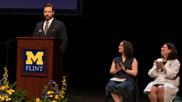Keith Saylor, in a suit, speaks at a lectern on stage. Two UM-Flint faculty members are in view seated on stage. They are clapping and looking at Keith.
