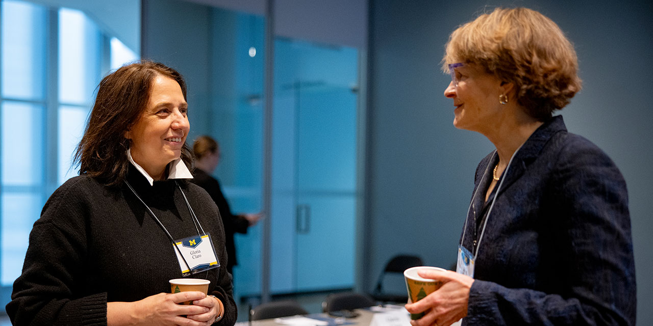 Two women conversing at a professional event, each holding a coffee cup and wearing conference lanyards.