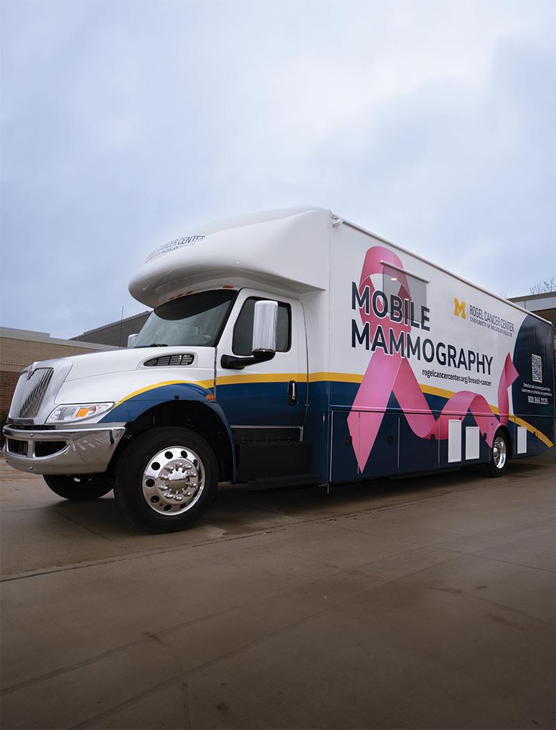 A parked mobile medical unit that reads “Mobile Mammography” and also features the pink breast cancer ribbon on the outside as well as a blue and yellow bottom border.