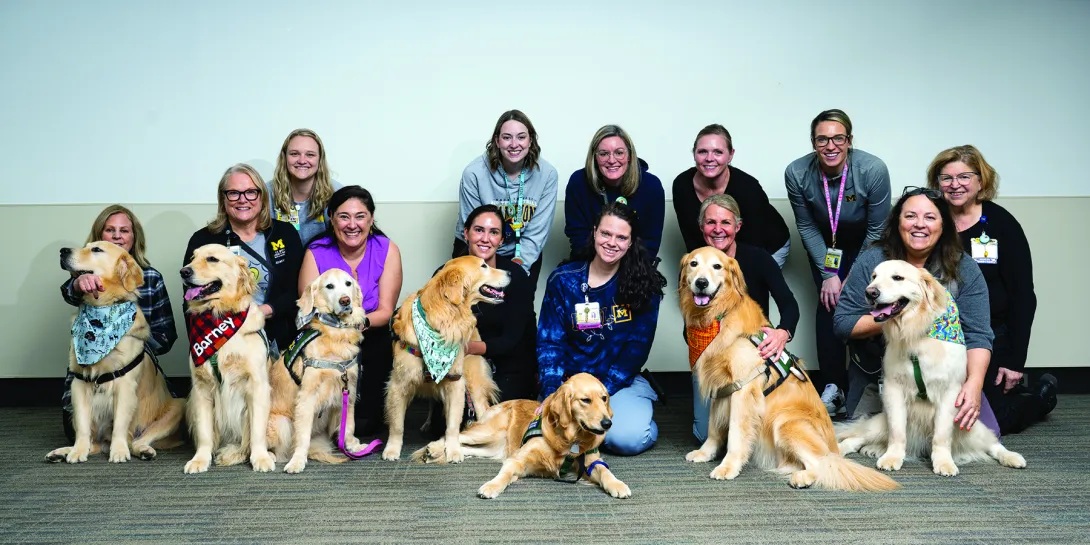 13 hospital workers pose with seven golden retrievers who work at the hospital. The retrievers are all wearing different colored bandanas. Everyone is looking at the camera and smiling.