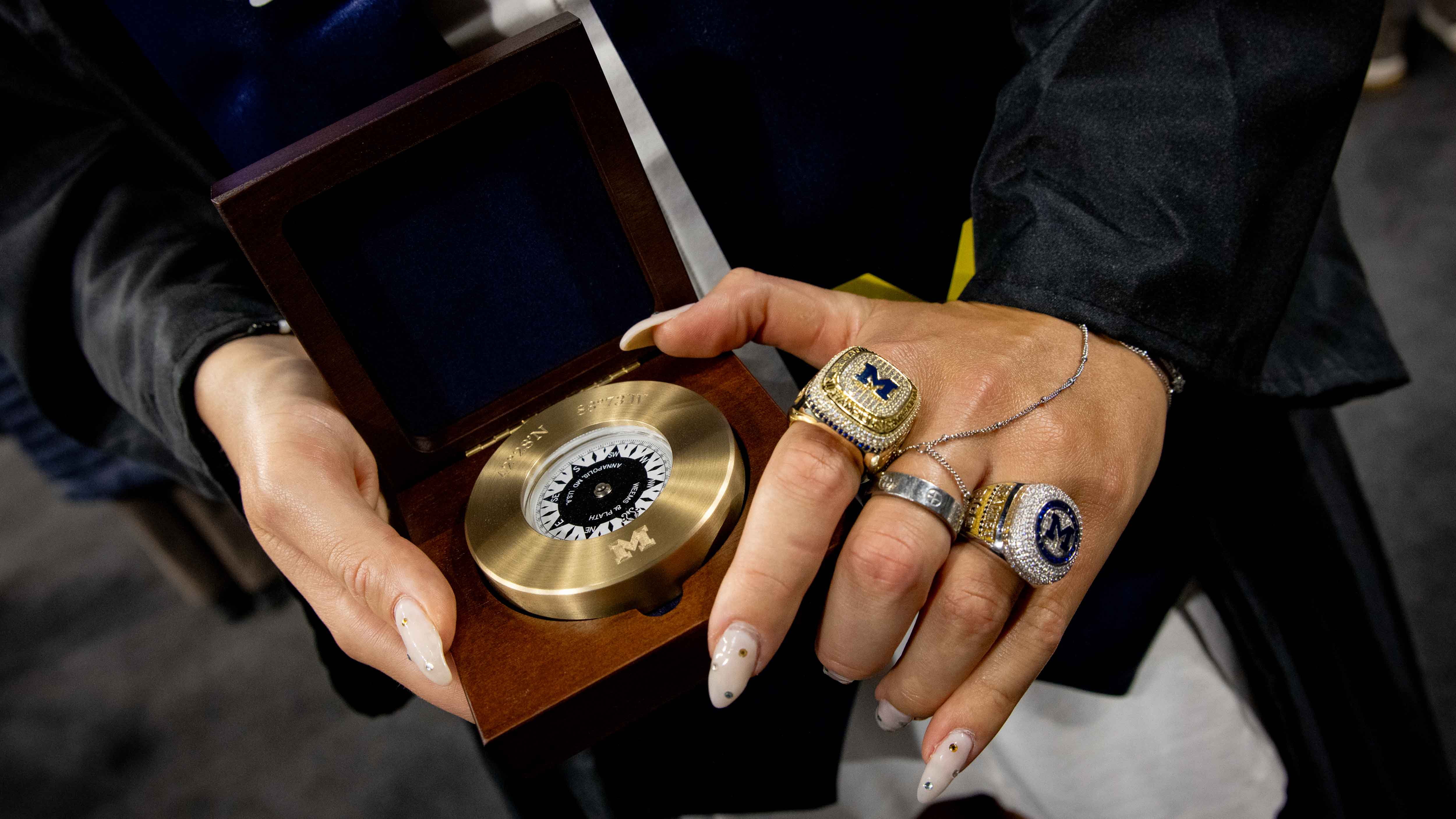 A close-up of a student-athlete graduate’s hands holding her golden compass, which is engraved with a Block M and placed in a wooden box. The graduate is wearing two large athletic rings on her left hand.