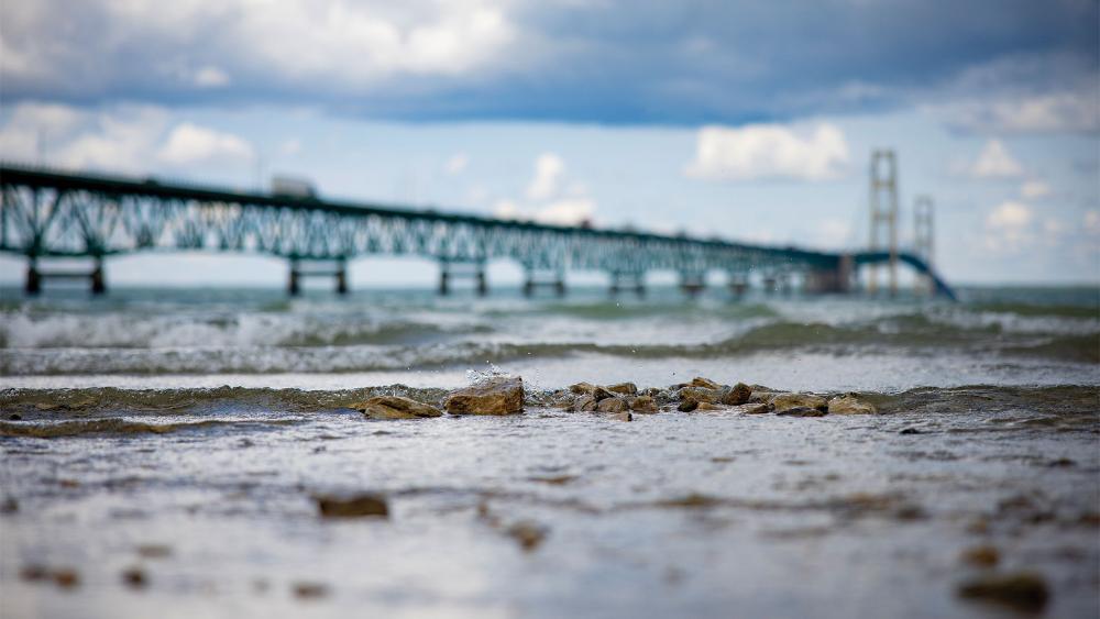 A rocky shore with waves lapping against it is in focus. A large bridge is in the background. The sky is cloudy.