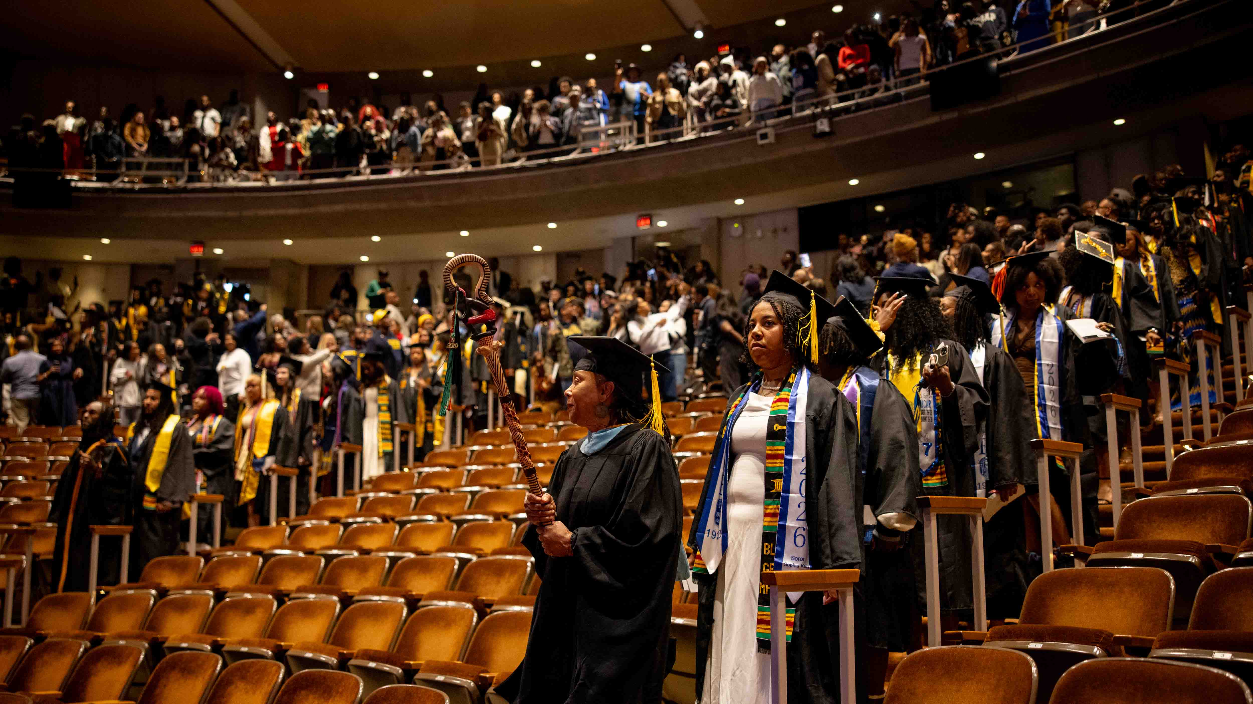 Graduates in caps and gowns entering the Power Center auditorium, led by a faculty member carrying a staff.