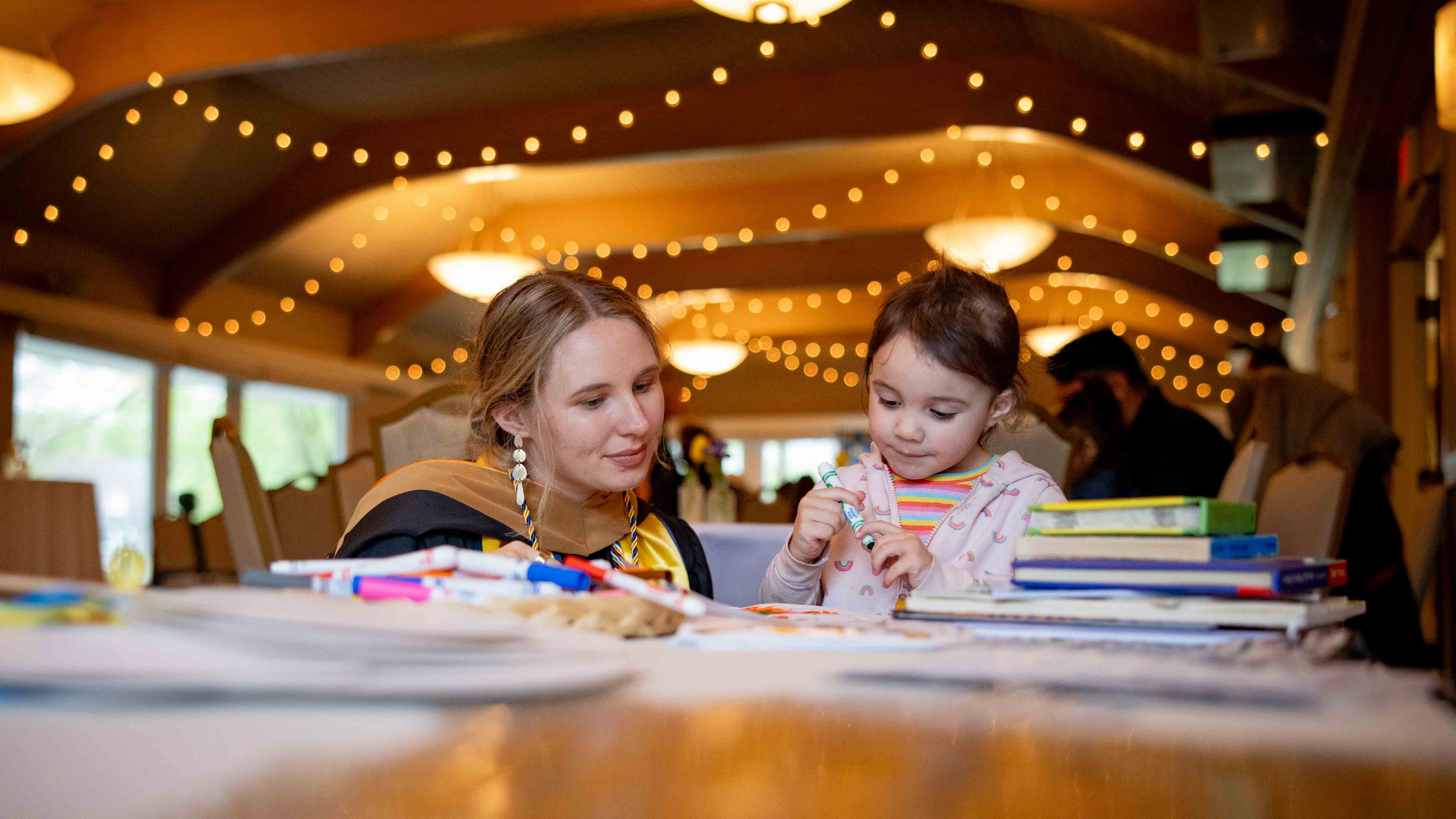 A graduate and her daughter sit at a table stacked with markers and books at the CEW+ Graduation Celebration. The graduate’s daughter is holding a marker and both are looking at a piece of paper.