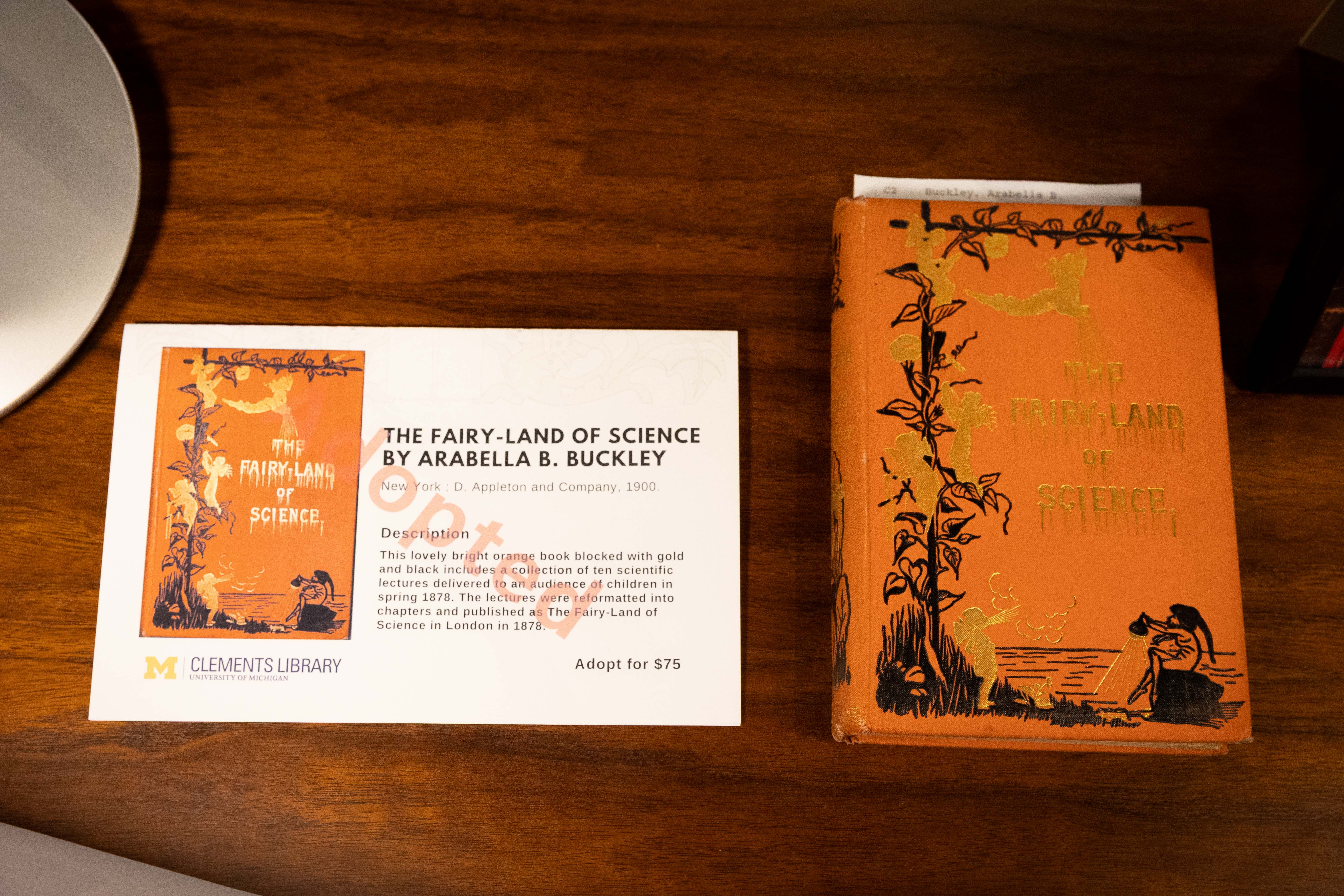 A vibrant orange copy of 'The Fairy-Land of Science' by Arabella B. Buckley sits beside an informational Clements Library card.