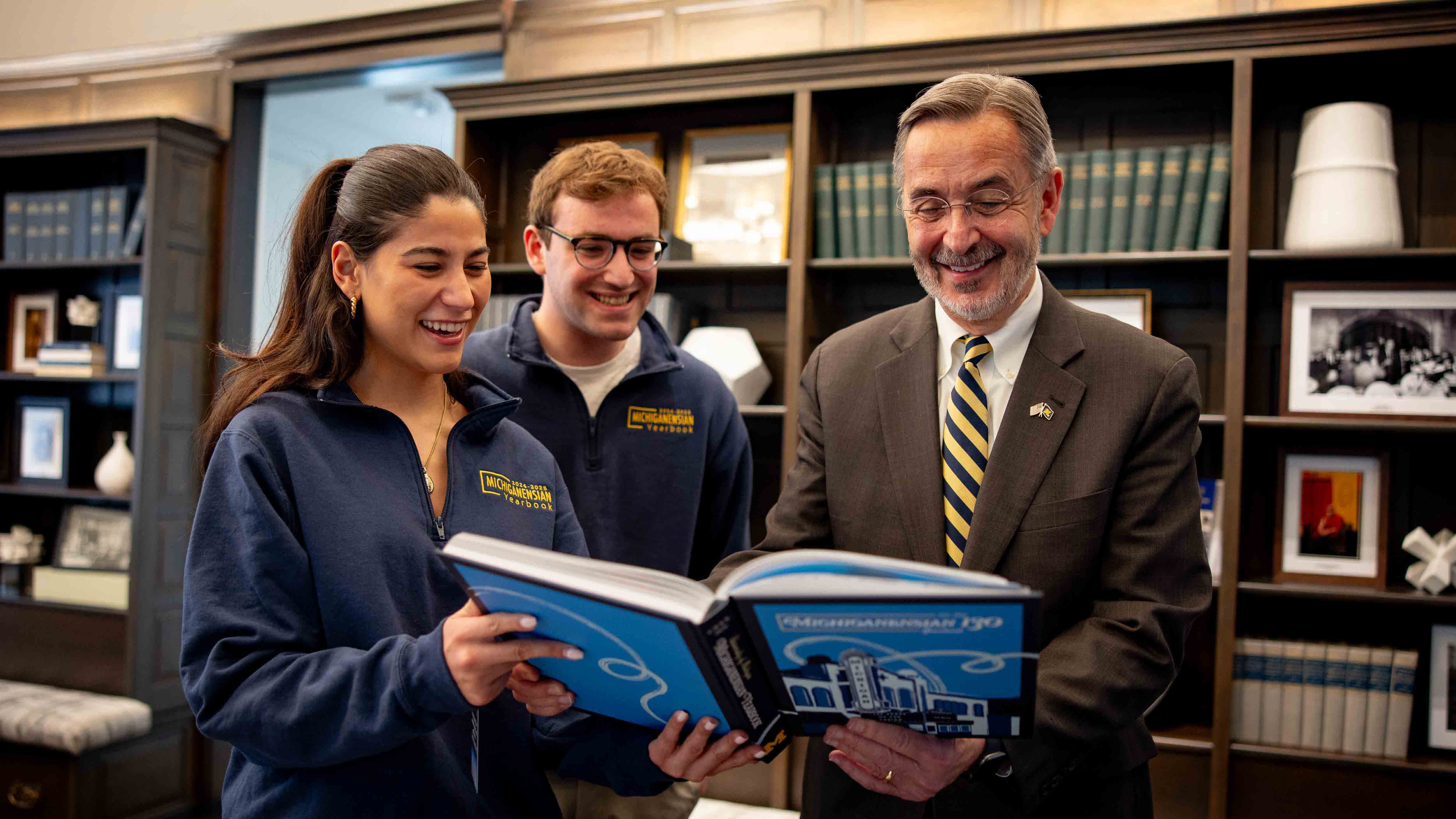  U-M students Gabriella Noble and Michael Reifman stand next to President Domenico Grasso. Noble and President Grasso hold open the 2025–2026 Michiganensian Yearbook, and all three are looking at it, smiling.