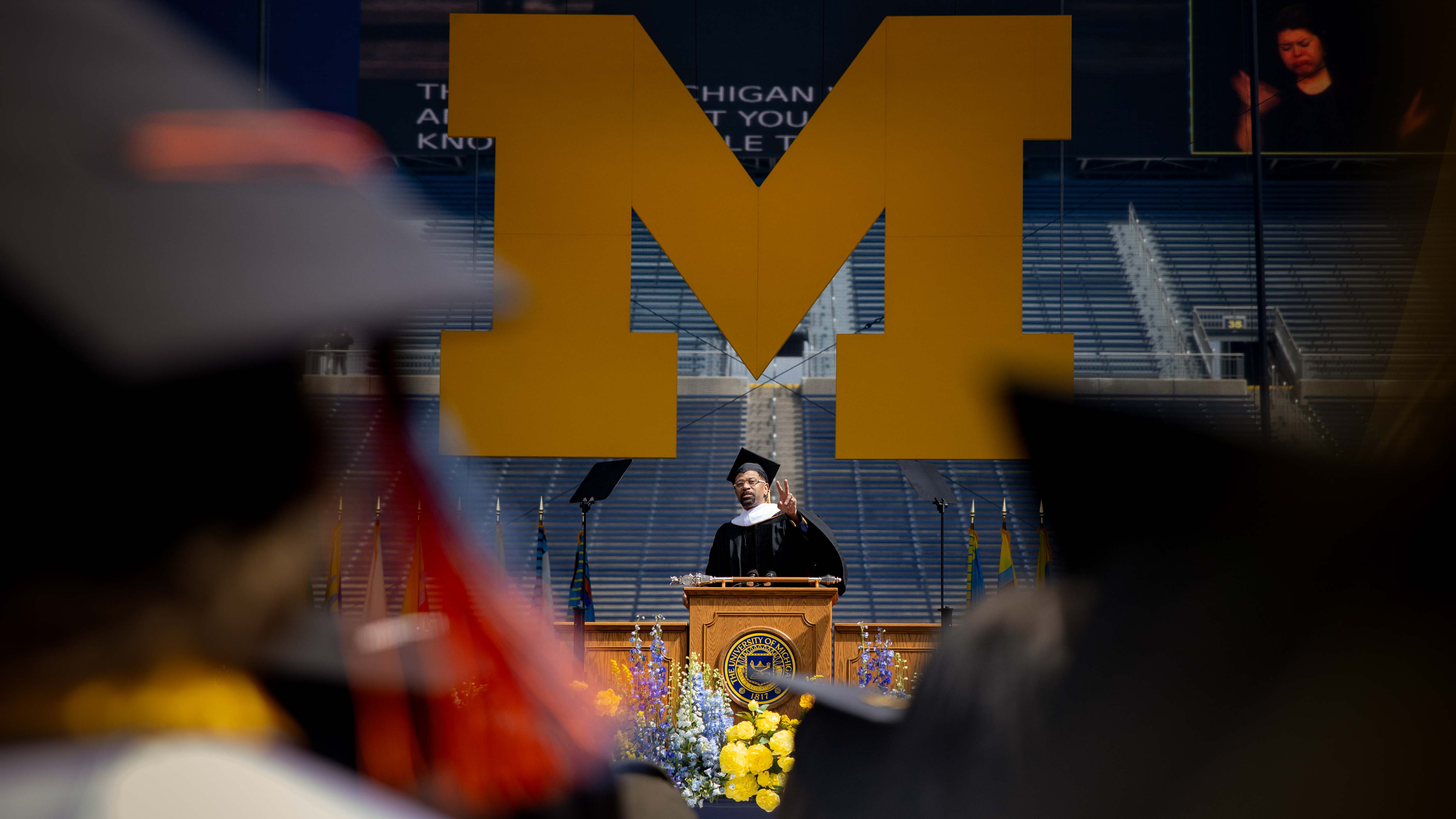 Jalen Rose speaks behind a podium, under a large Block M, at the Big House.