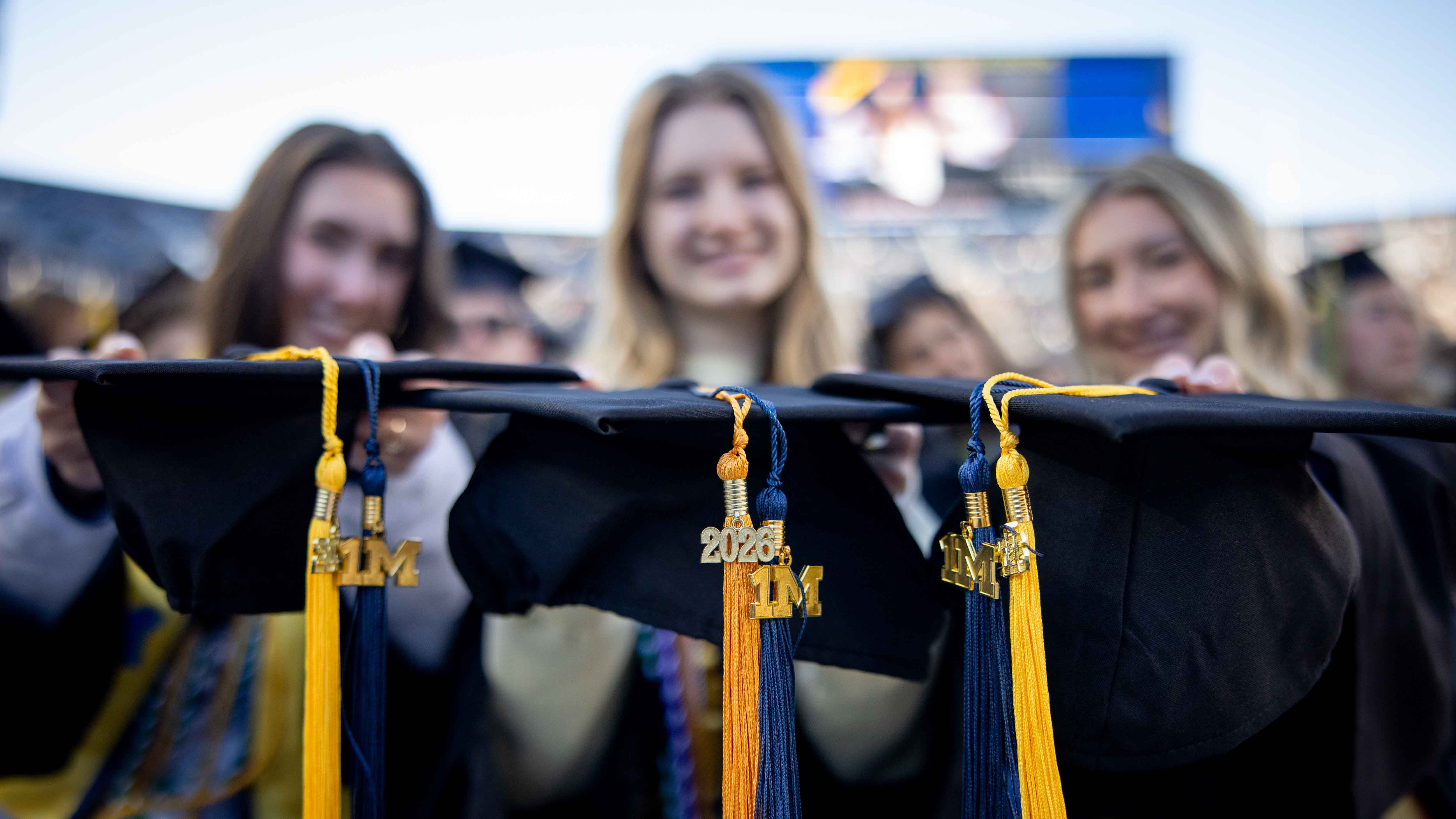 Three female students out of focus hold their three graduation caps up to the camera, in focus. The caps have yellow 2026 tassels and blue 1 million tassels.