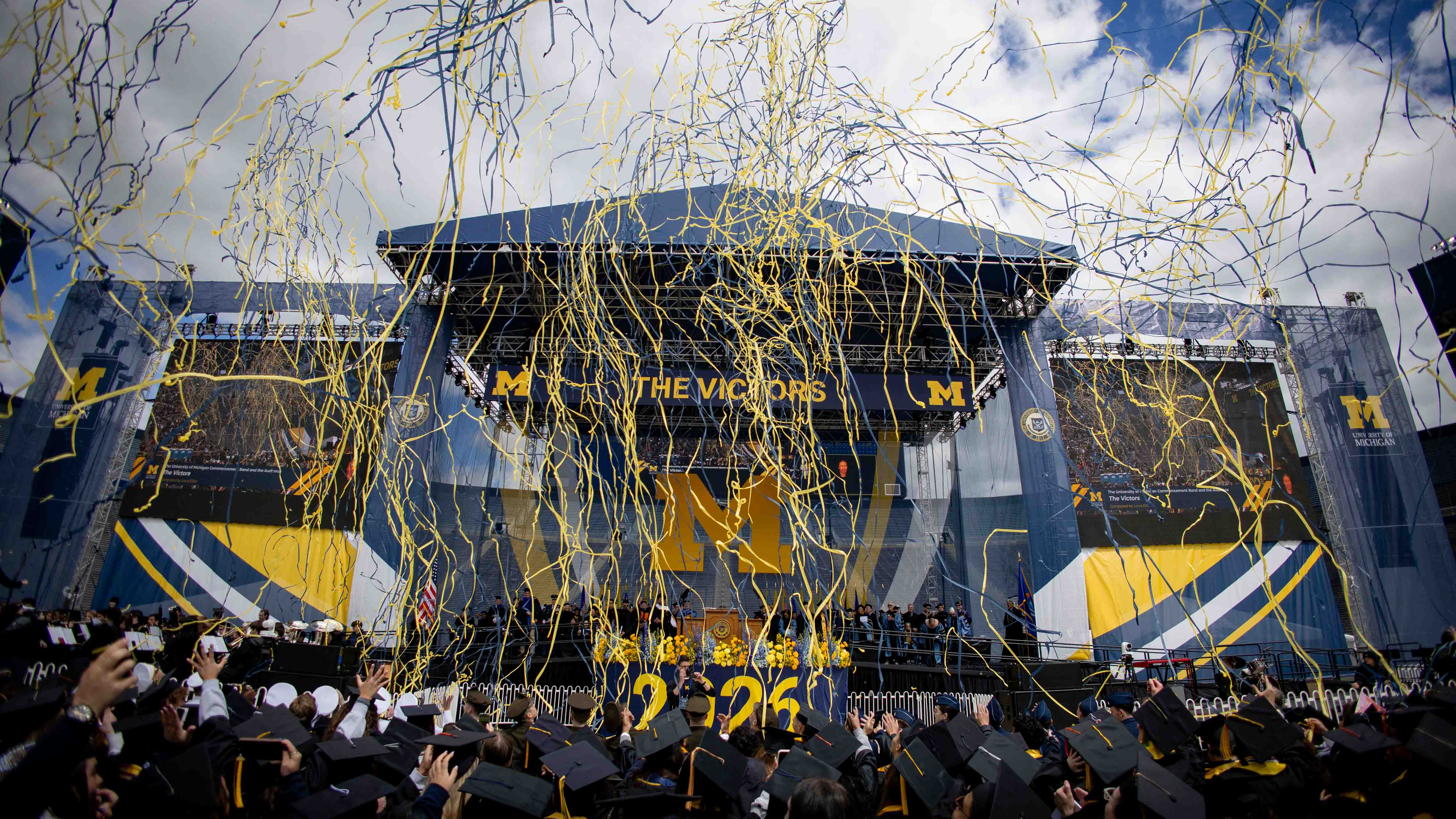  A crowd of students in graduation caps and gowns cheering with yellow and blue streamers in the air, in front of a stage decorated with Block Ms, the numbers 2026, 
and the words The Victors.