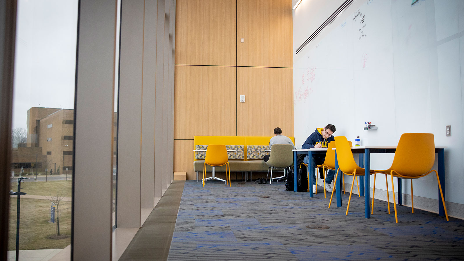 Students study in a space with a wall of windows and colorful furniture.