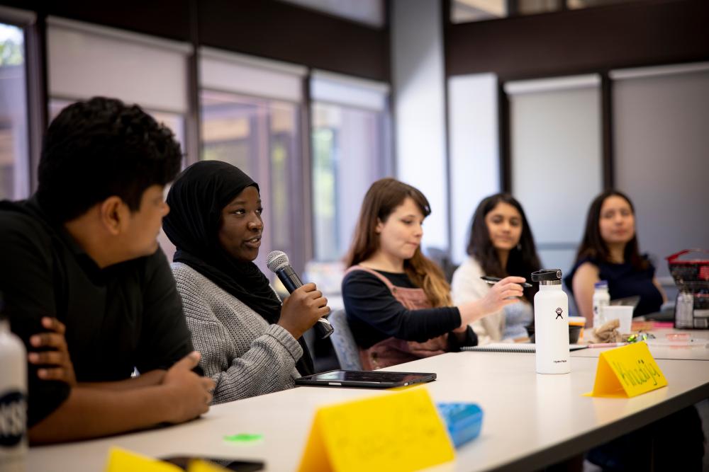 Students sitting at a conference table. One is speaking with a microphone while others listen and take notes.