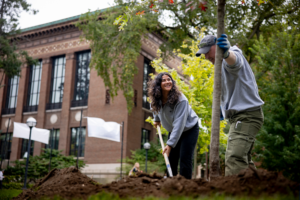 Two people with shovels smiling as they dig into the ground to plant trees in front of the library.