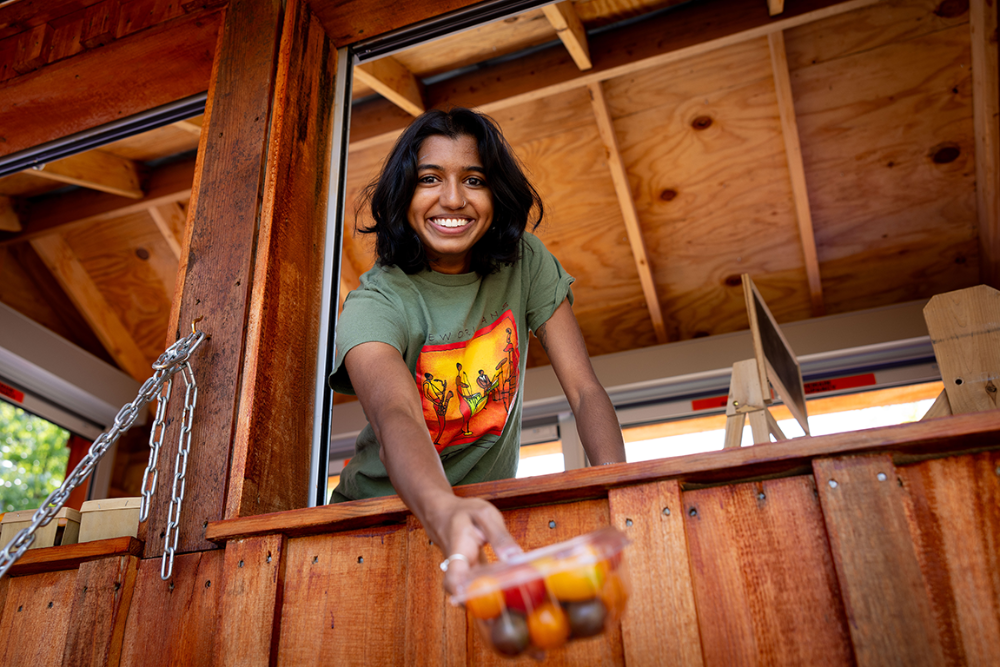 A student smiling reaching out with a plastic box of tomatoes at a wooden farm stand.