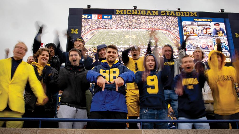 A group of students in the stands at a U-M football game cheering, dressed in maize and blue, in front of the jumbotron with a picture of the stadium.