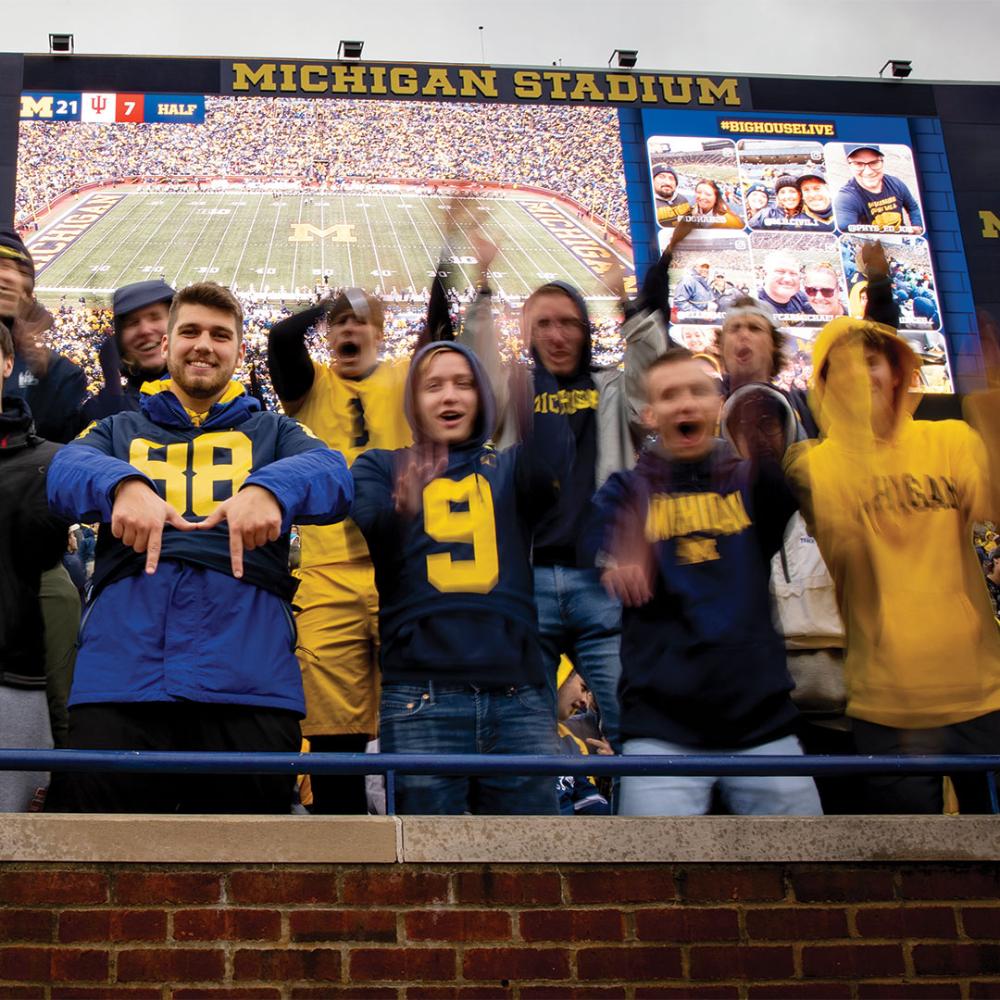 A group of students in the stands at a U-M football game cheering, dressed in maize and blue, in front of the jumbotron with a picture of the stadium.