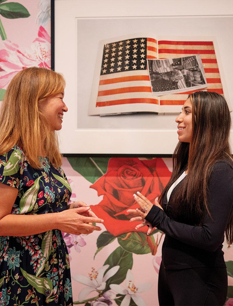 Two women smiling and talking to each other in front of a framed photo of an American flag with a black and white photo on top of it.