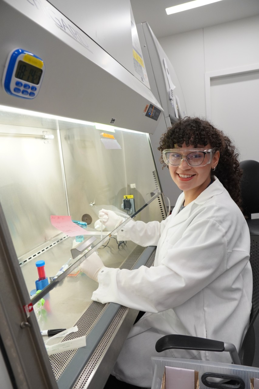 Sydney Richardson works at a biosafety cabinet in a lab. She is wearing a white lab coat, clear safety goggles, and white latex gloves, and is handling a small sample or slide inside the fume hood. Various lab supplies, including tubes and containers, are visible inside the cabinet. A blue digital timer is mounted on the hood above. She is turned toward the camera, smiling.