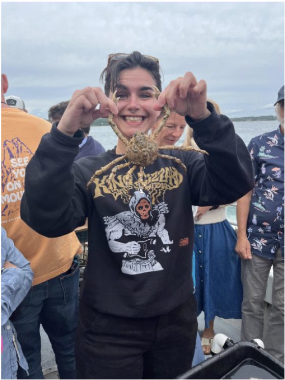 Ever O'Donnell, with a big smile, holds up a crab by its two front legs. They are wearing a dark sweatshirt and dark pants.
