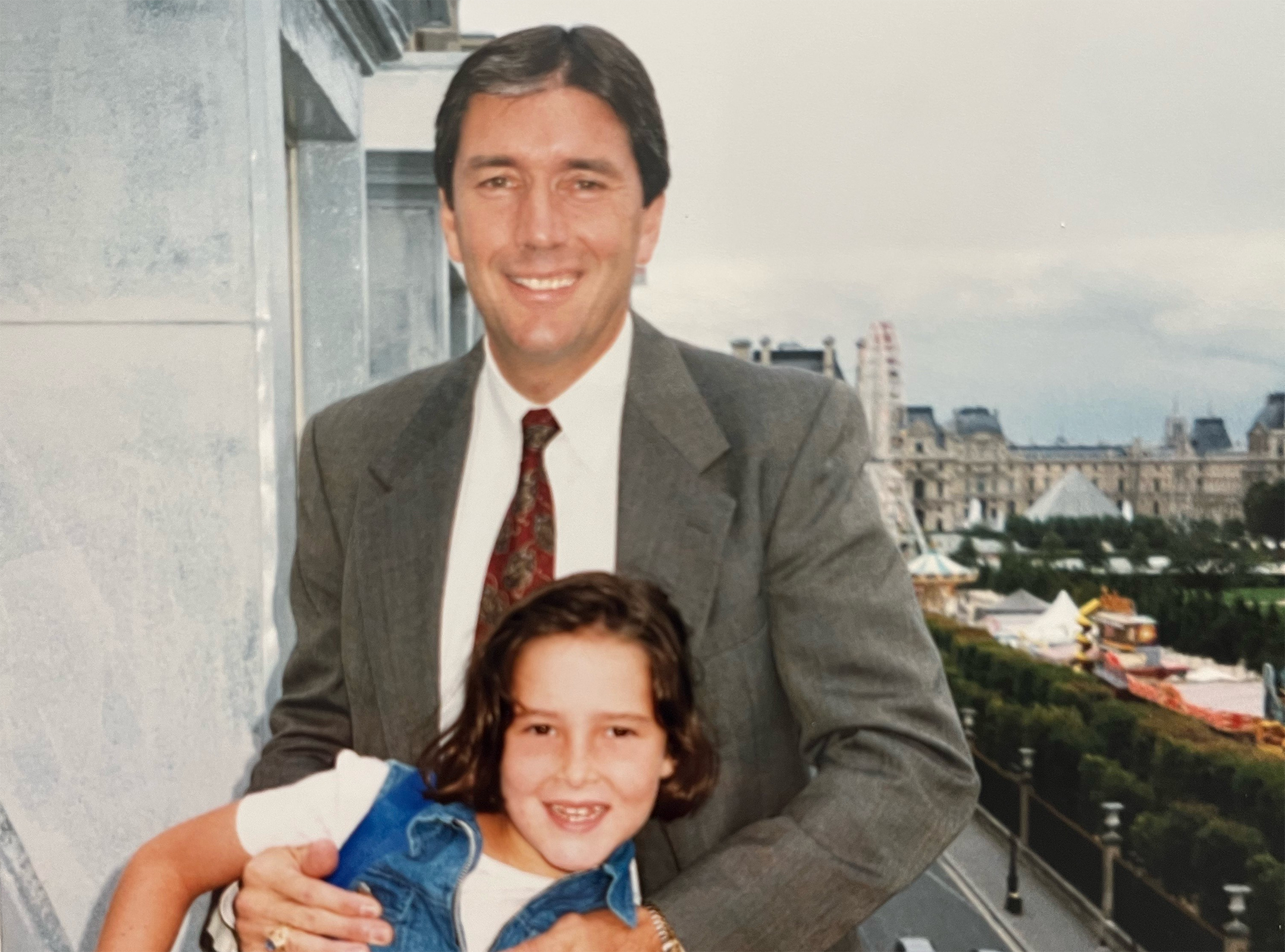 A young Laura Raines and her stepfather pose on a balcony against a backdrop of the Paris skyline.