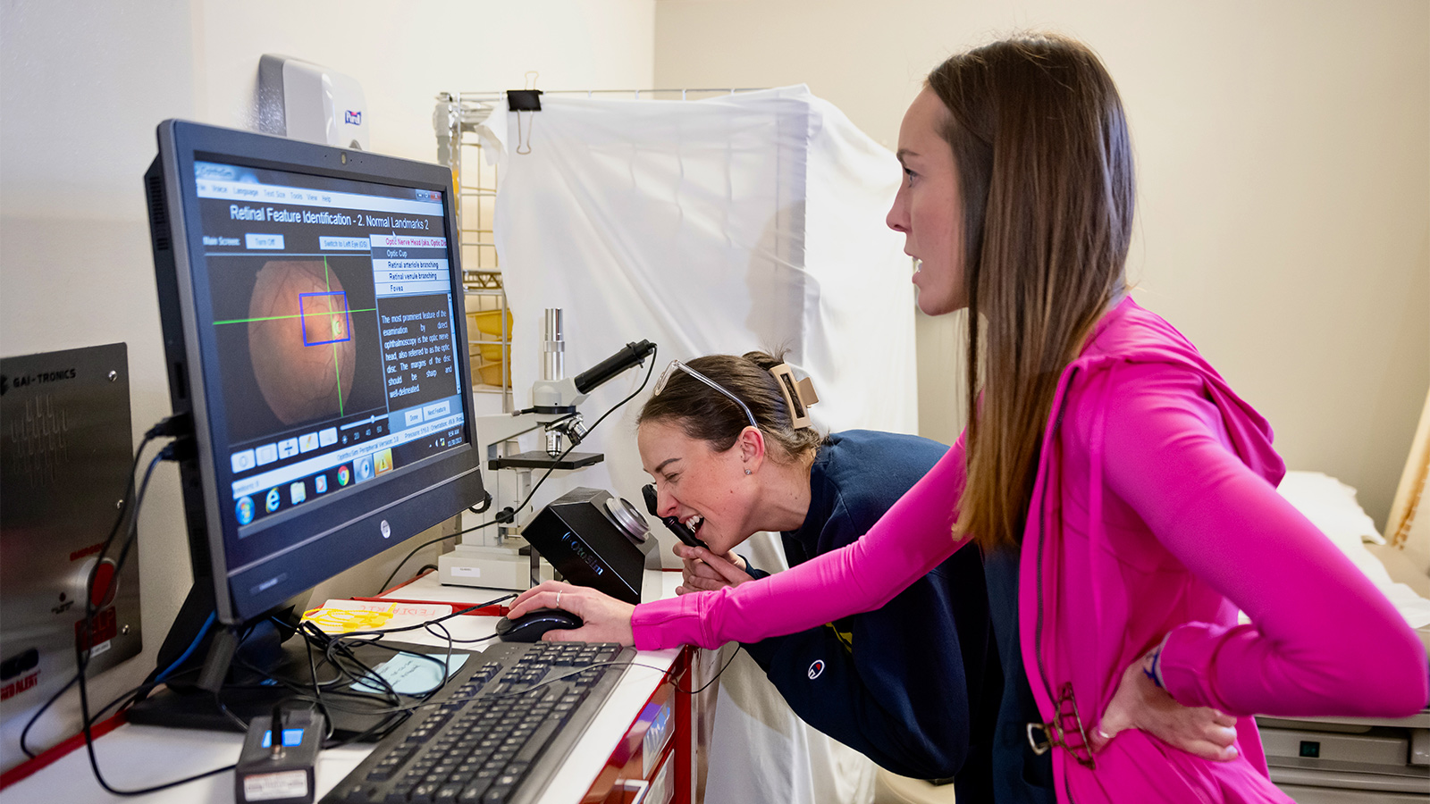 A nursing student looks at a computer monitor displaying retinal feature identification software, while another student uses specialized eye examination equipment. 