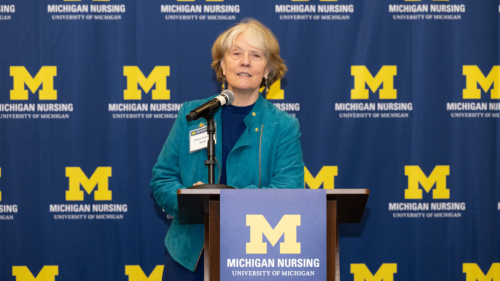 Dean Patricia Hurn speaks at a podium featuring a Michigan Nursing banner, set against a matching backdrop.